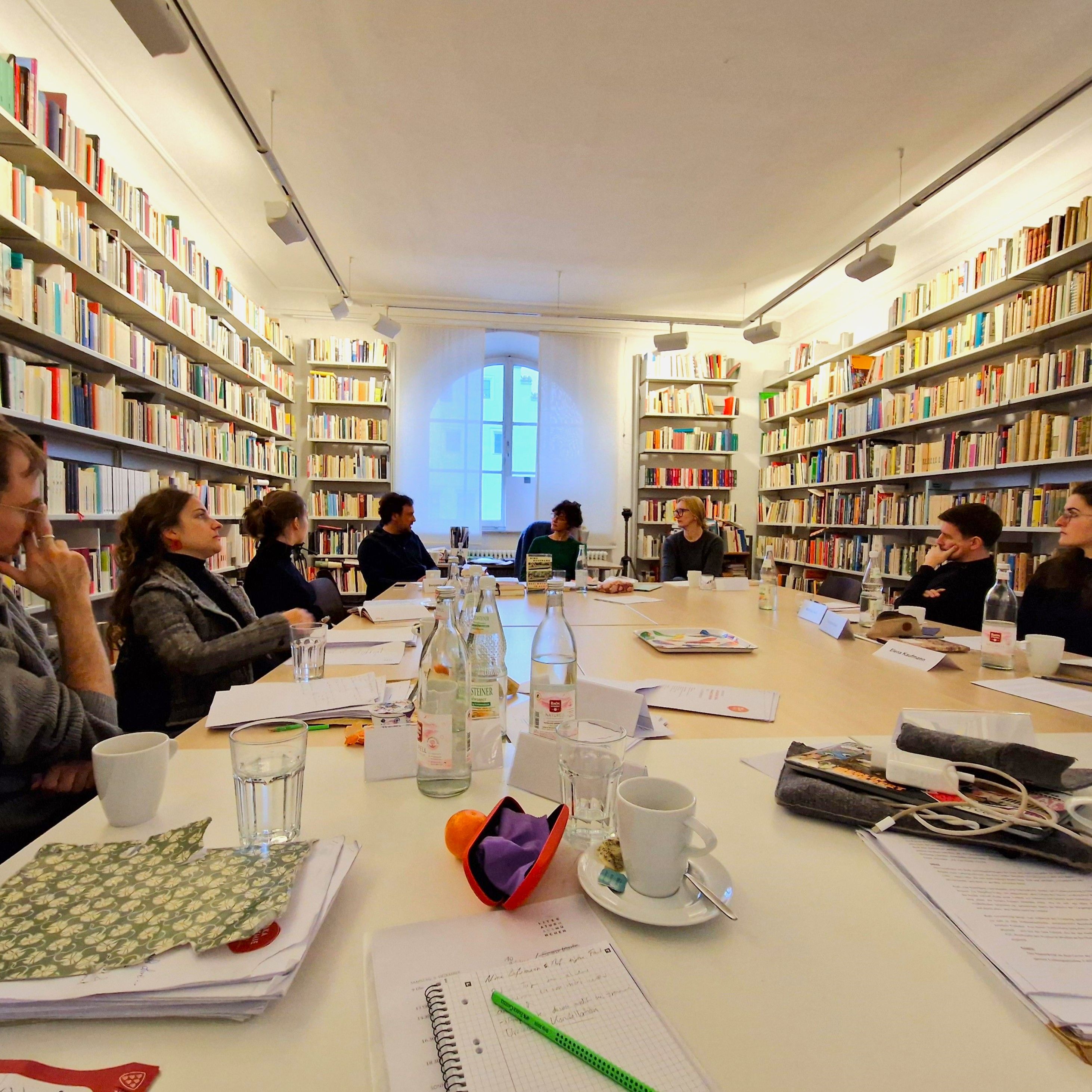 Frauen und Männer sitzen um einen großen Tisch mit Papier, Wassergläsern und Büchern.