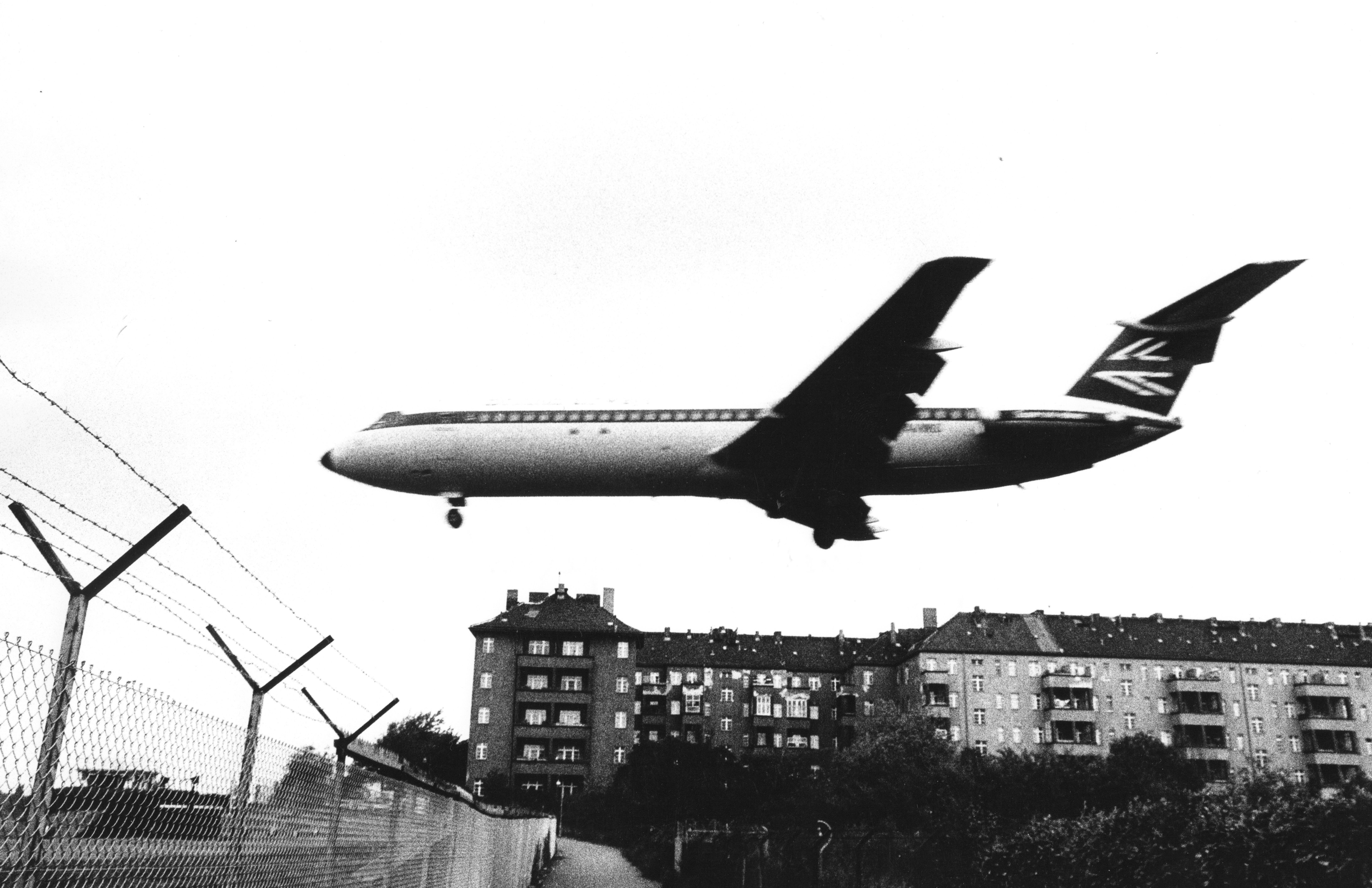 Schwarz-weiß-Foto mit einem sehr tief fliegendes Passagierflugzeug über Wohngebäuden und einem Stacheldrahtzaun in Berlin-Tempelhof